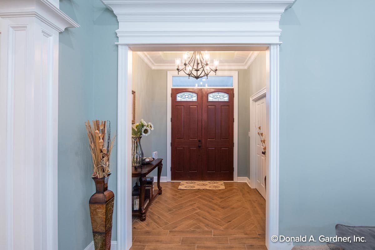 Foyer view of double wooden doors with glass transoms, herringbone wood floor, and ornate chandelier.