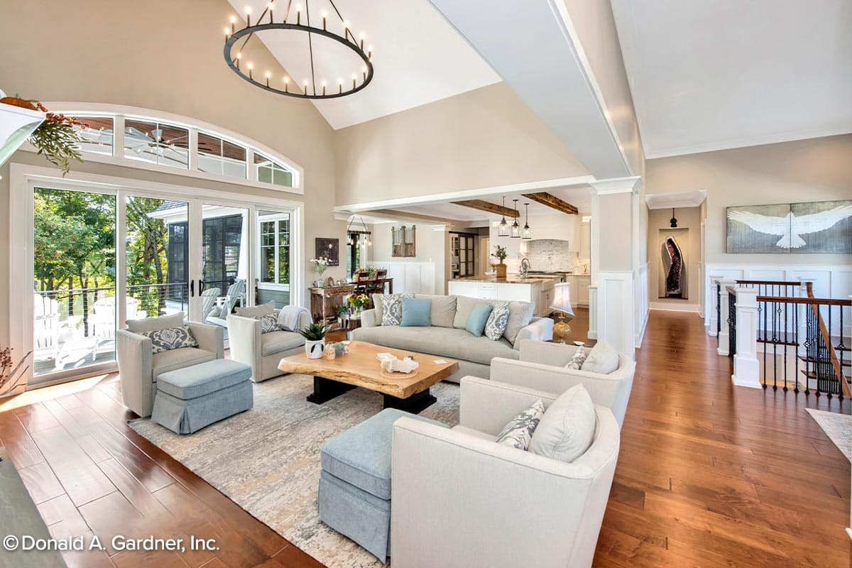 Interior view of a great room with vaulted ceiling, large windows, open kitchen, and a staircase.