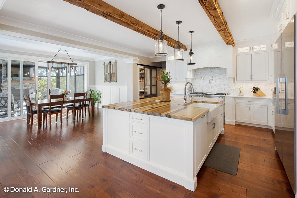 Interior view of a kitchen with a large island, farmhouse sink, white cabinets, and rustic wood beams on the ceiling.