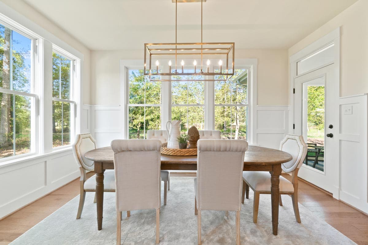 Dining room with wood table, upholstered chairs, rectangular chandelier, and large windows with view of trees.