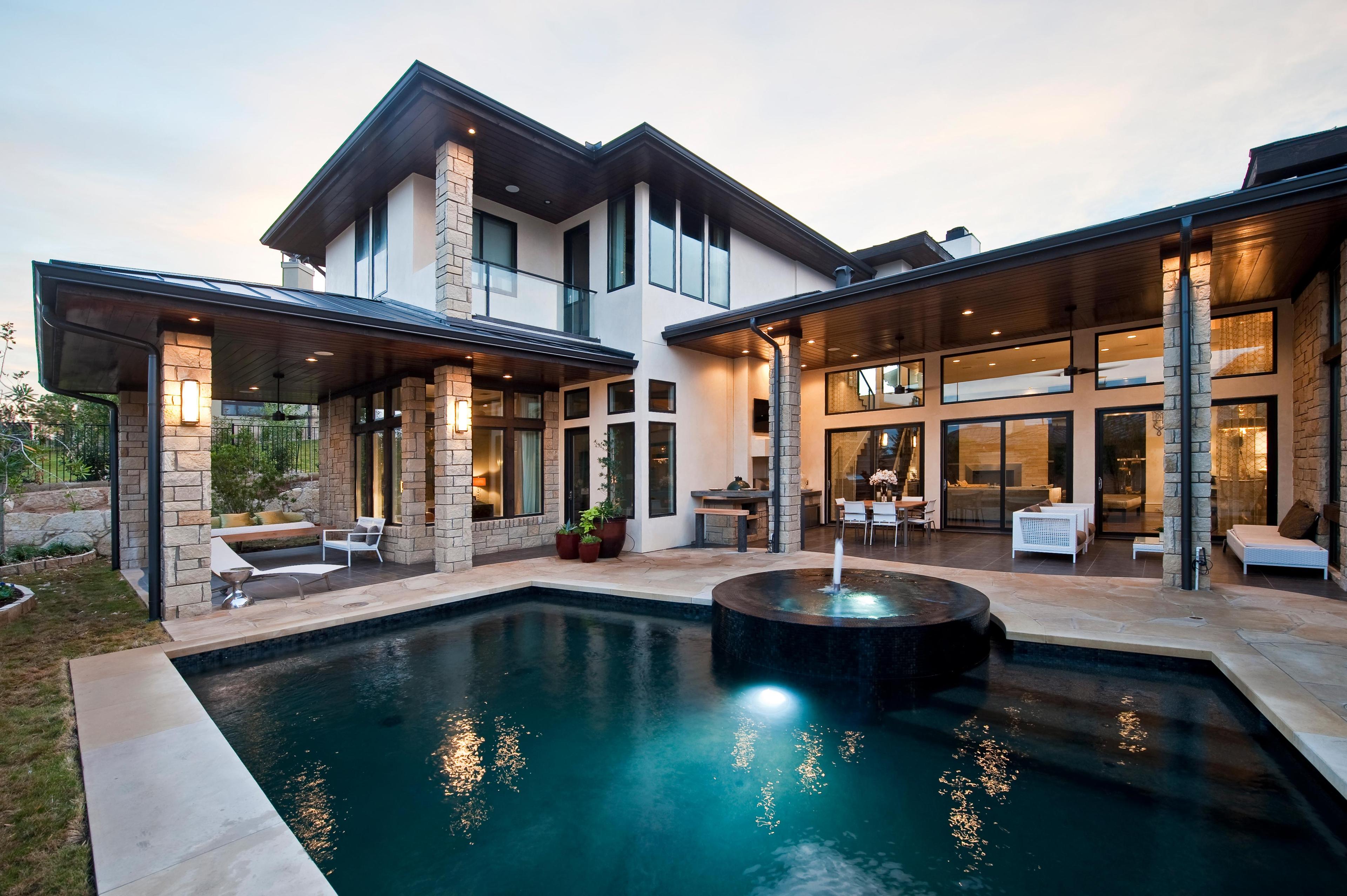 Two-story modern house exterior with expansive windows, covered patios, stone columns, and a swimming pool with a circular fountain.