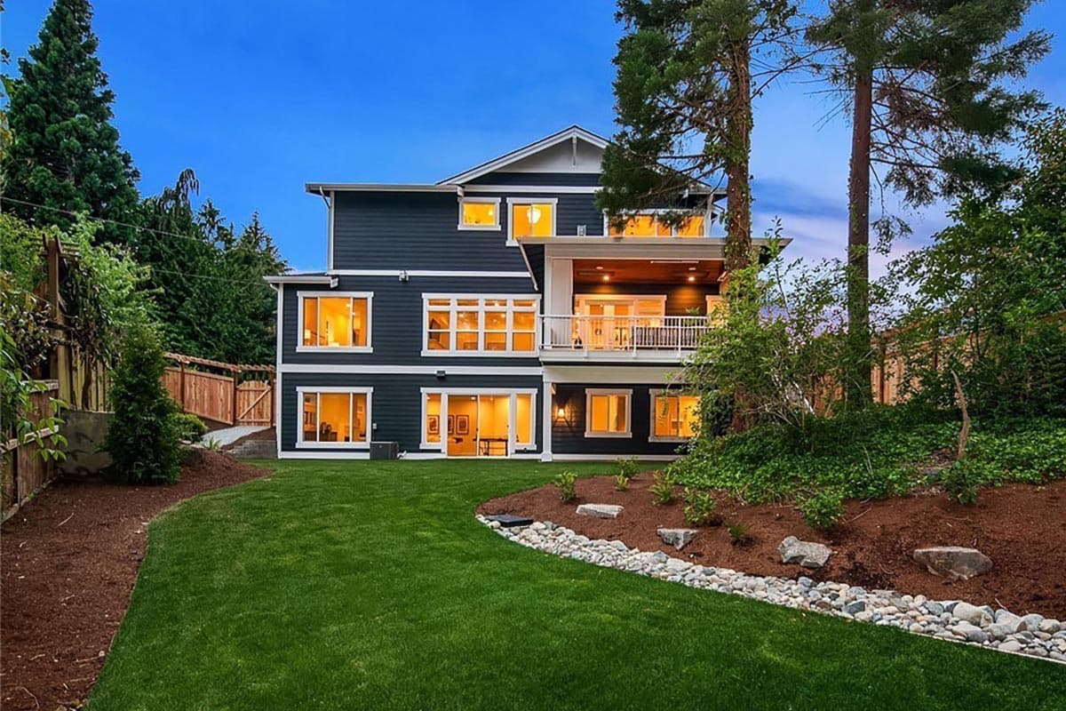 Modern house plan exterior, three stories, dark siding, white trim, large windows, covered balcony, sloped backyard.