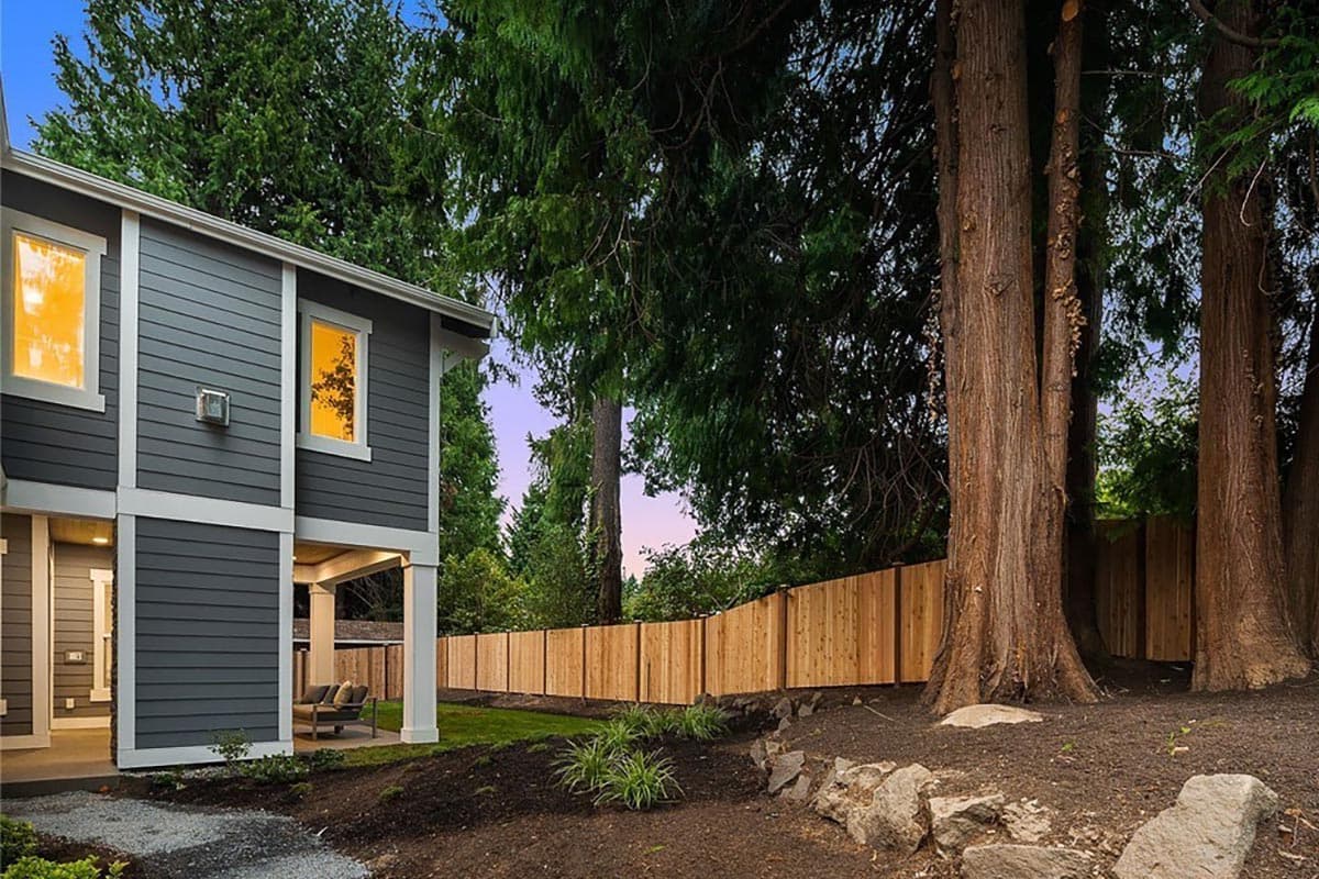 Two-story house exterior with dark gray siding, white trim, covered porch with seating, and large trees.