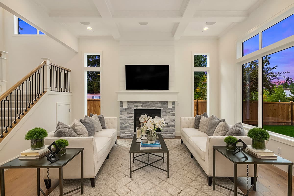 Great room with coffered ceiling, stone fireplace, and large windows. Staircase with metal railing is visible.