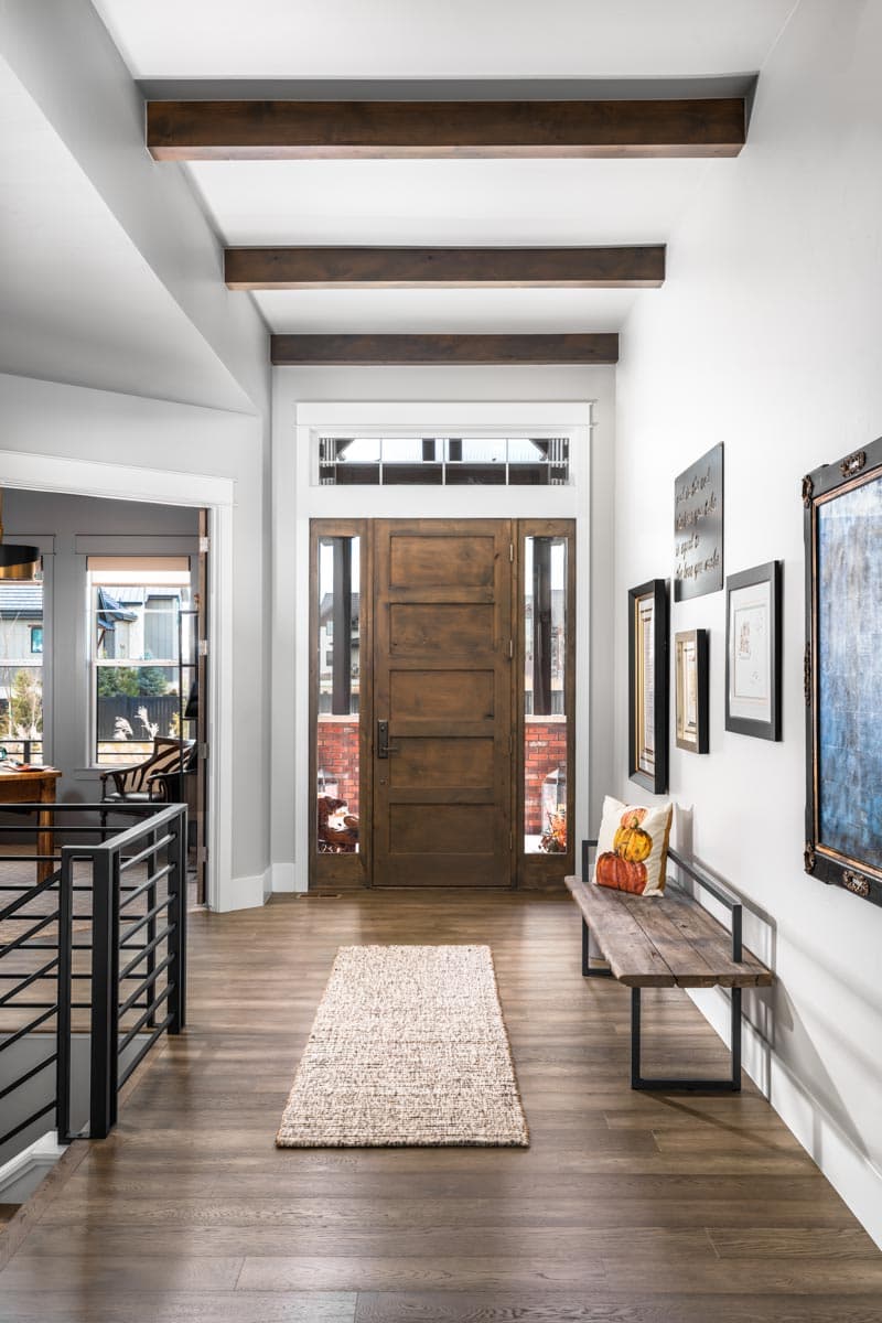 Interior view of a house entryway featuring a wooden double door with sidelights, exposed wood ceiling beams, and a hallway with a modern railing.
