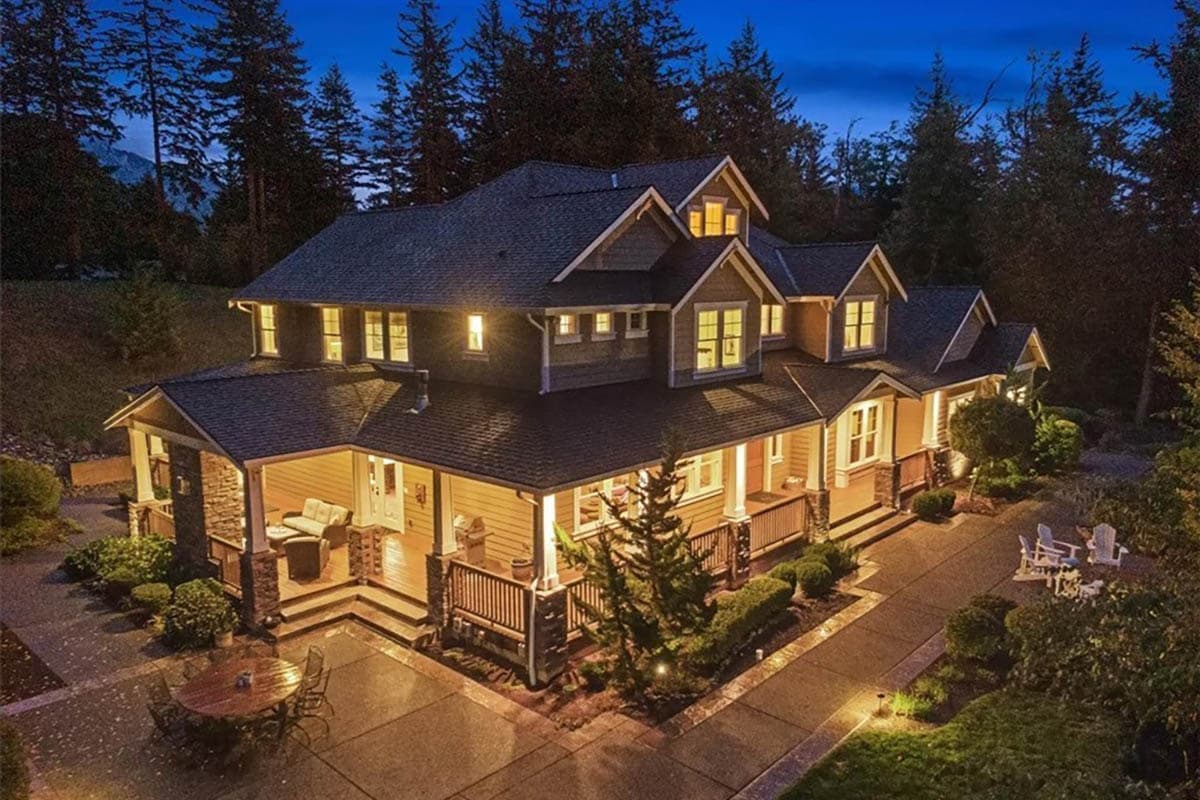Two-story house plan exterior with gabled dormers, wide wraparound porch with stone columns, and multi-level rooflines.