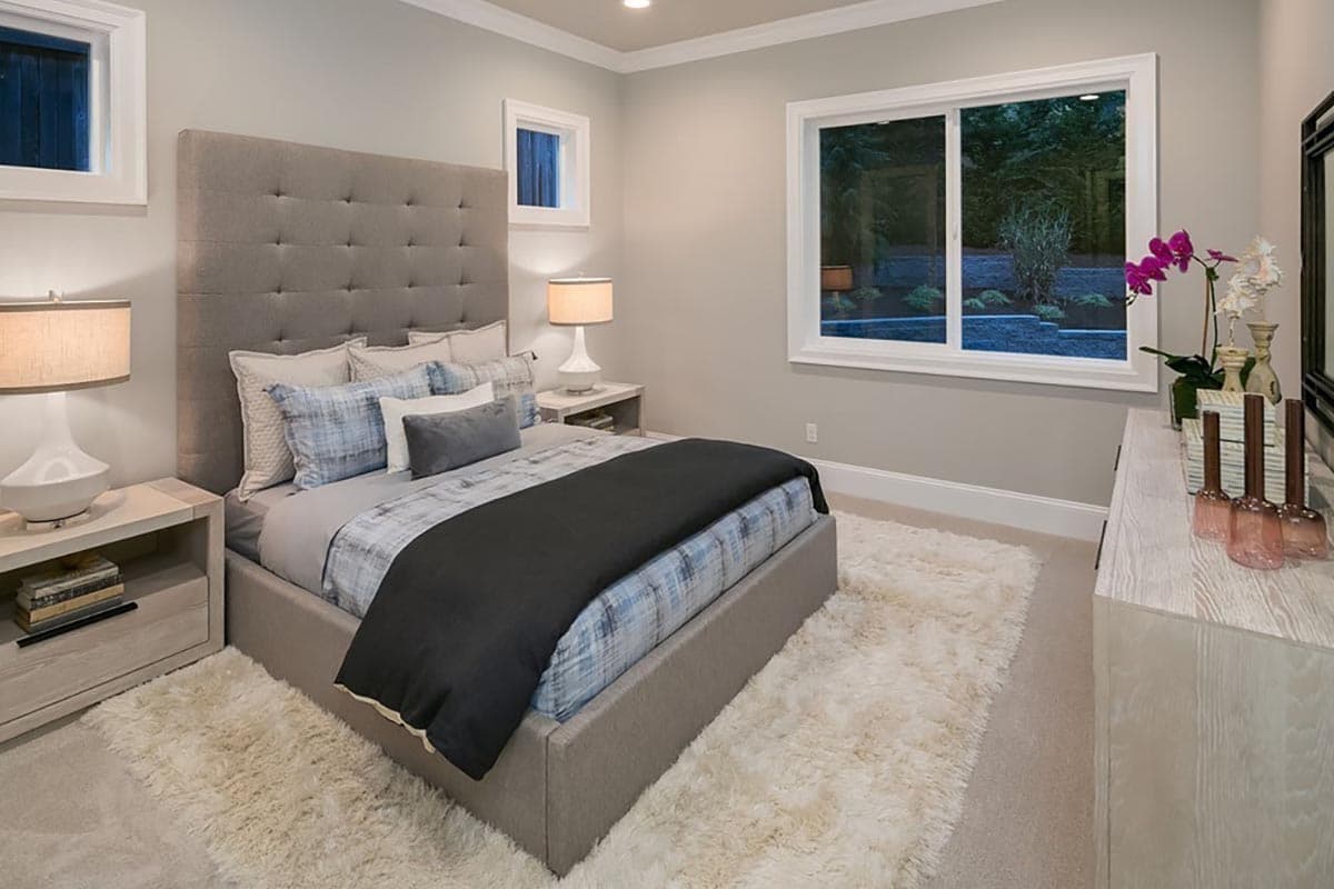 Bedroom interior with upholstered headboard, queen bed, dresser, and large picture window overlooking landscaping.