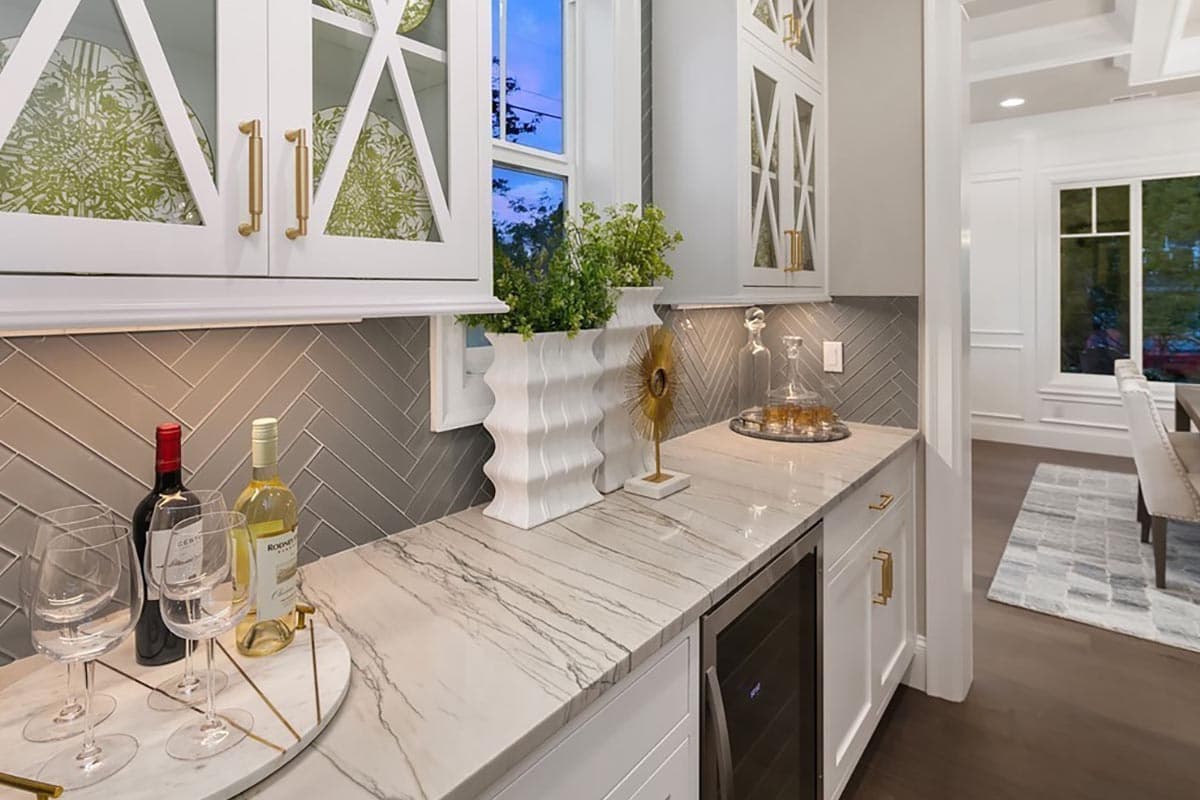 Interior view of a kitchen bar with white cabinets, marble countertop, herringbone tile backsplash, wine cooler, and glass-front display cabinets.