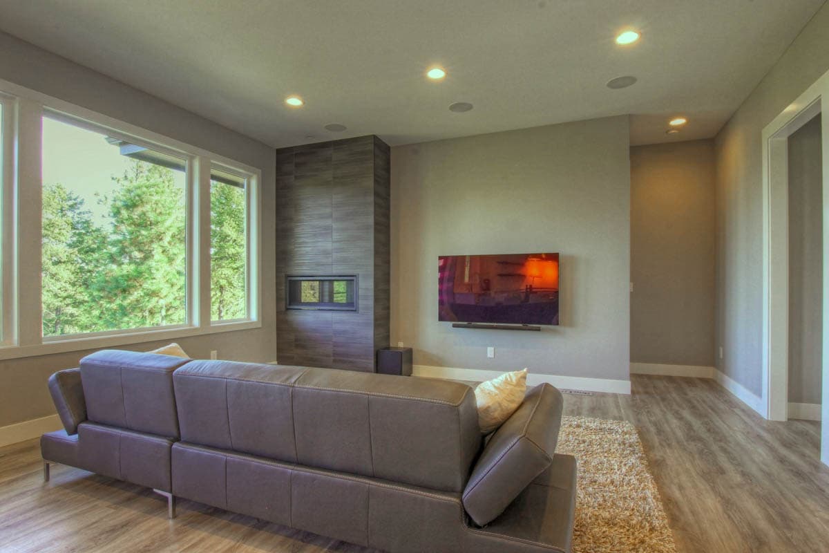 Living room with leather sofa, fireplace, and large windows overlooking trees. Wall-mounted TV.