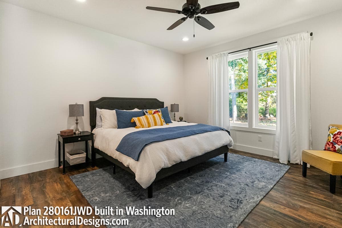 Interior view of a bedroom with a bed, nightstand, ceiling fan, and large windows with white curtains.