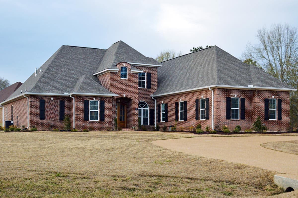 House plan exterior: Red brick, two-story home with a hip roof, arched entryway, dormer window, and attached side garage.