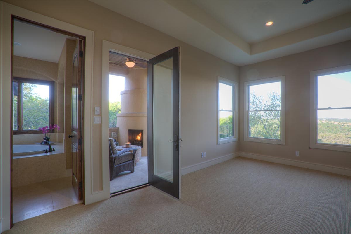 Bedroom with tray ceiling, multiple windows, and doorway to a bathroom and outdoor patio with fireplace.