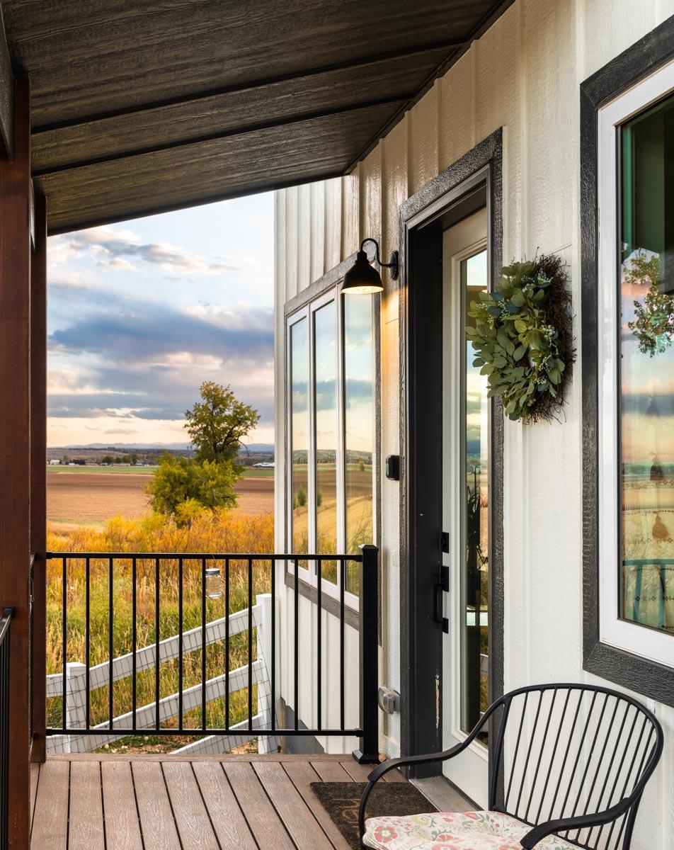 Modern farmhouse exterior with covered porch, vertical siding, large windows, and metal railings overlooking a rural landscape.