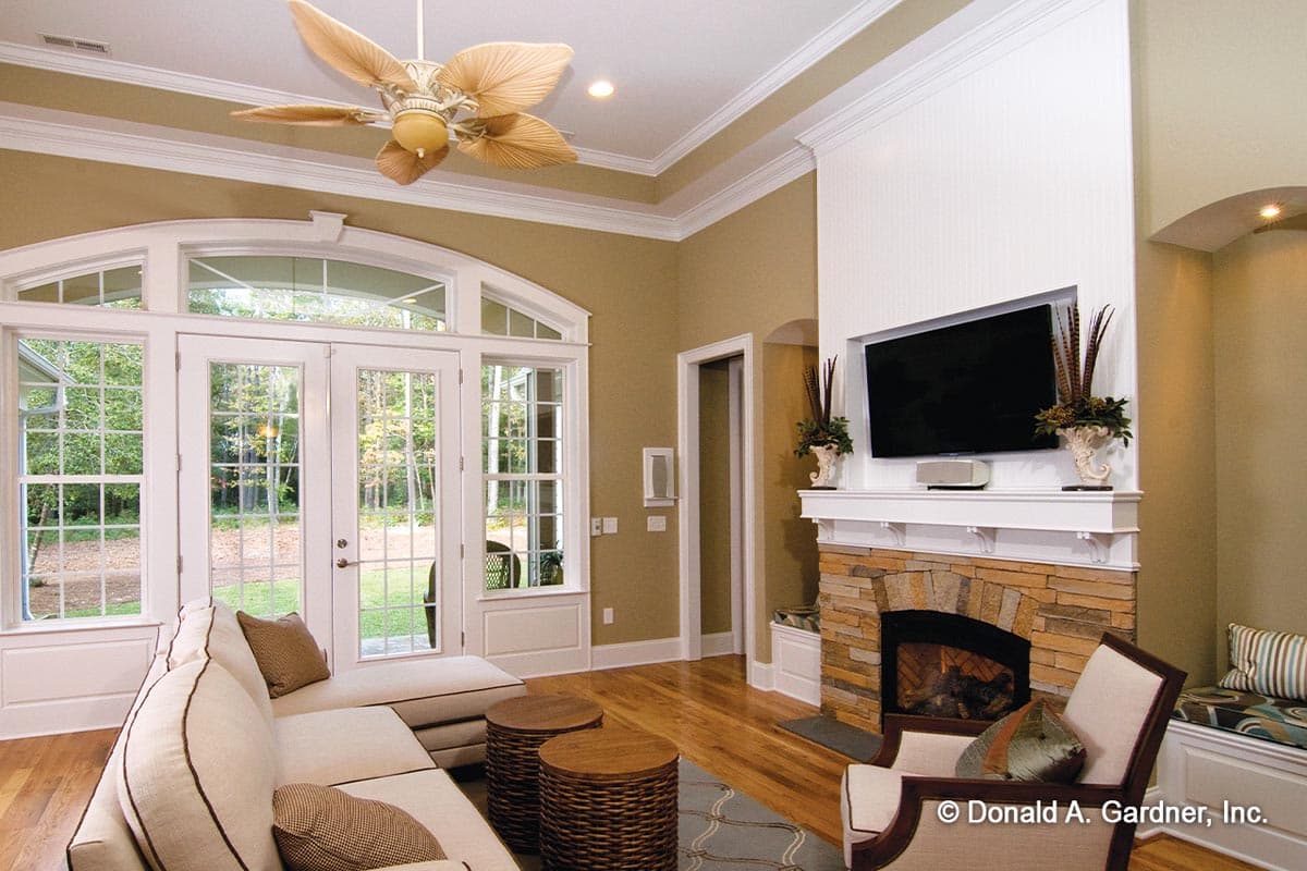 Interior view of a living room with a large sectional sofa, stone fireplace, and wall-mounted TV, featuring large windows and double doors.