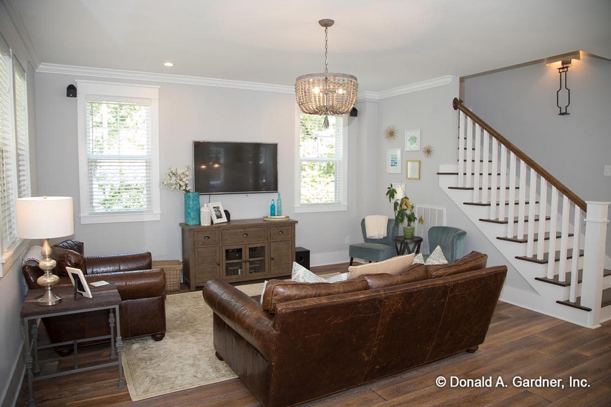 Living room interior with leather sofas, media console, chandelier, and white spindle staircase.