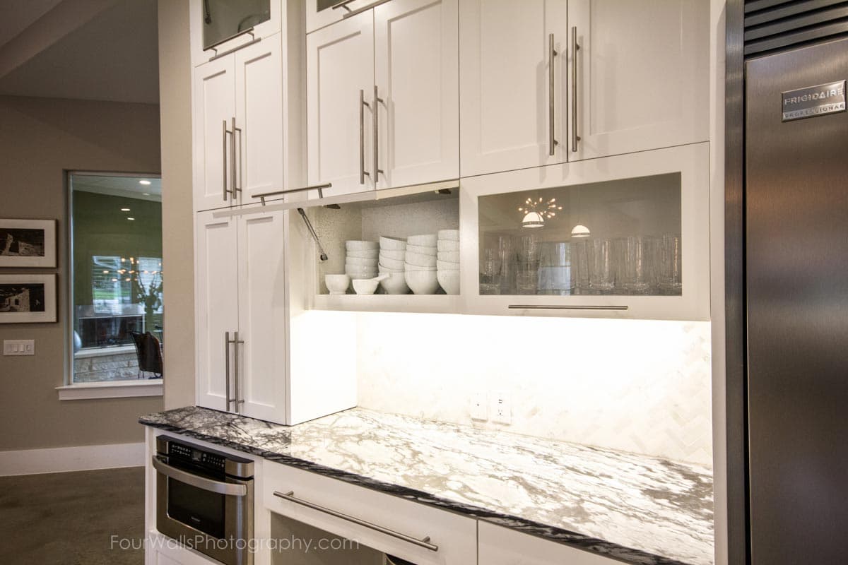 Kitchen cabinetry with built-in microwave, marble countertop, glass-fronted upper cabinets displaying glassware, and open shelving with stacked bowls.