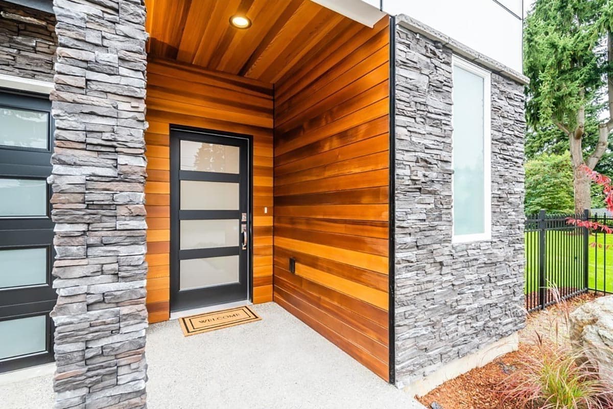 Modern house exterior with stacked stone pillars, wood siding, and a glass-paneled door.