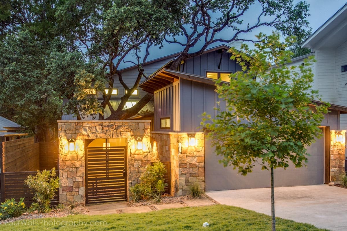 House plan exterior. Two-story house with a garage and stone wall with a gate. A tree grows through the house exterior.