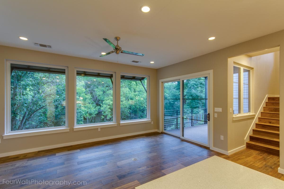 Interior view of a living area with three large windows, sliding glass door to deck, and wood staircase. Modern design.