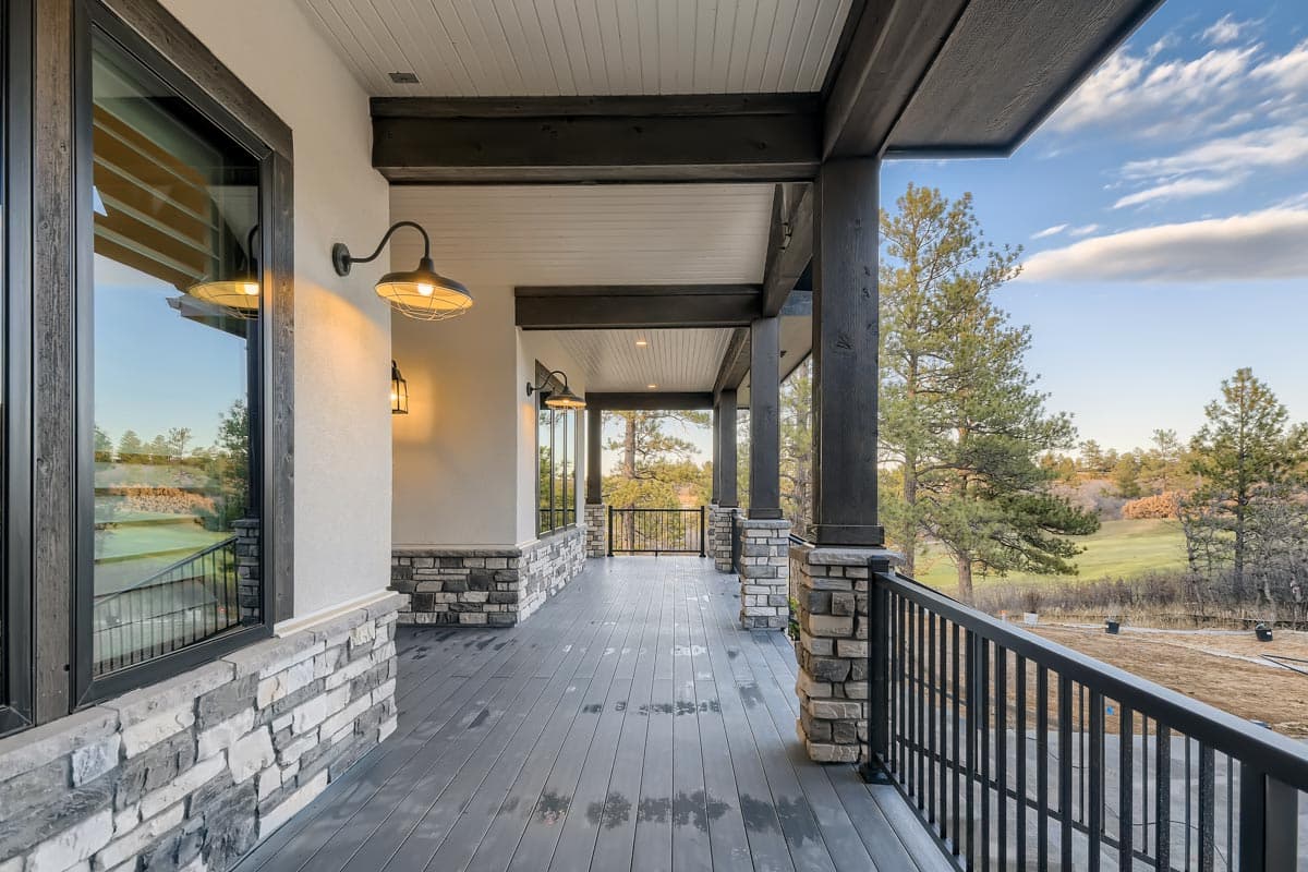 Covered porch with wood beams, stone accents, industrial sconces, and a railing overlooking a landscape.