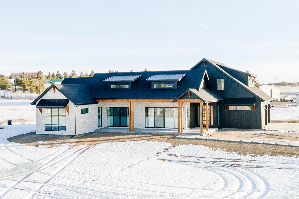 House plan exterior: Modern design with white stone and dark siding, gable roof, dormers, and covered porch with timber columns.