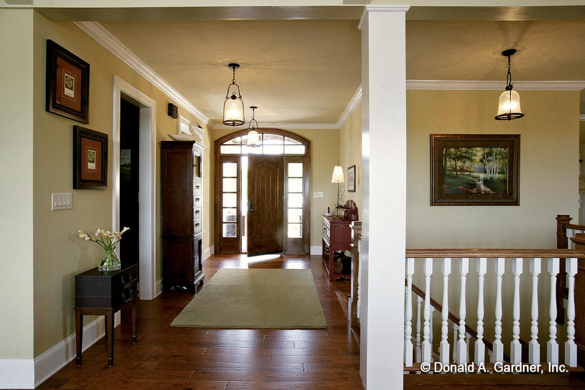Interior view of a wide foyer with double wood doors, arched transom, hardwood floors, and a staircase with white balusters.
