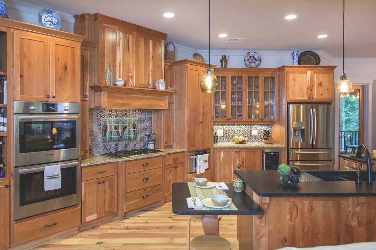 Kitchen with wooden cabinetry, double oven, gas range with decorative tile backsplash, island with sink, and stainless steel refrigerator.