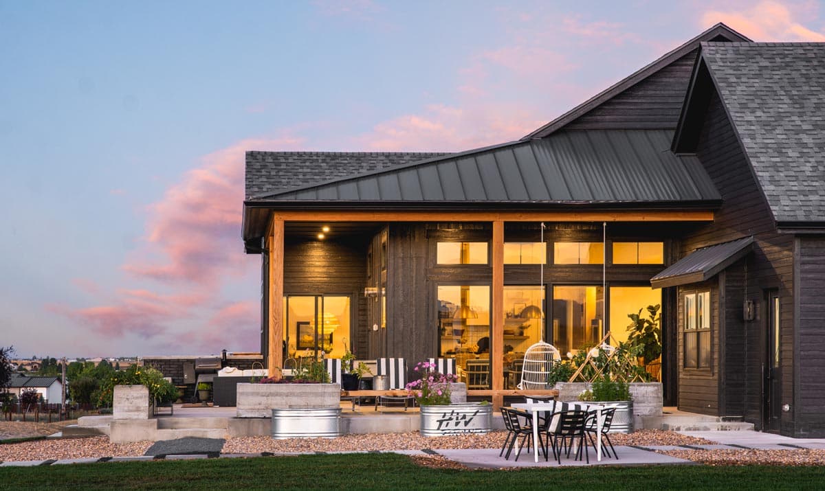 Modern Farmhouse exterior with dark wood siding, metal roof, covered patio with large windows, and outdoor seating area.