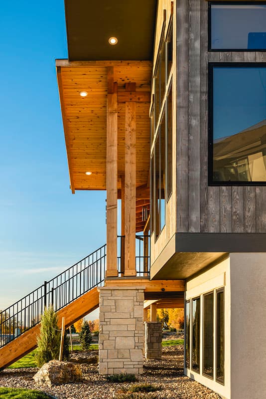 Modern house exterior showing wood siding, large windows, stone base, and timbered columns supporting an overhang with recessed lighting.