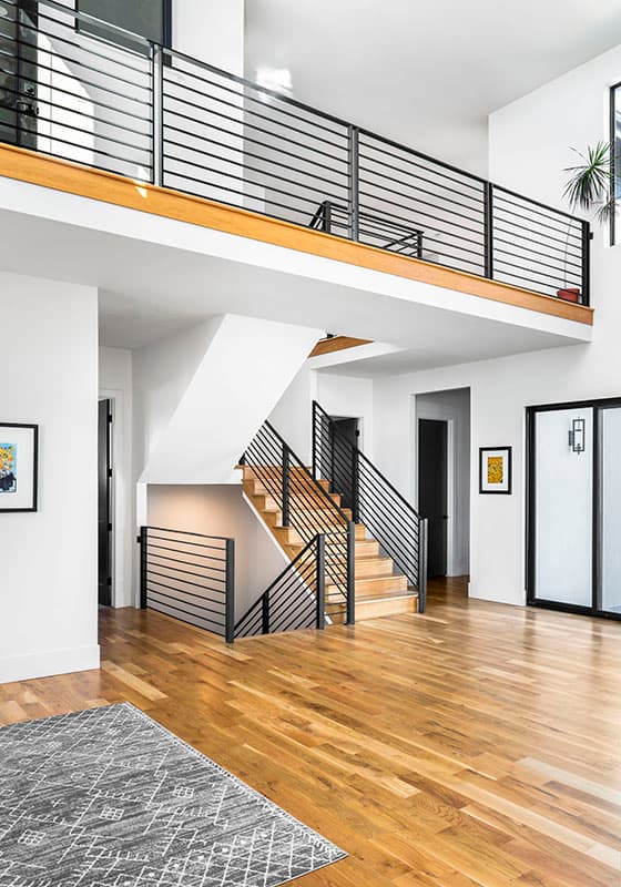Modern interior with open loft, wood staircase, and black horizontal railing. Wood floors and minimal decor visible.