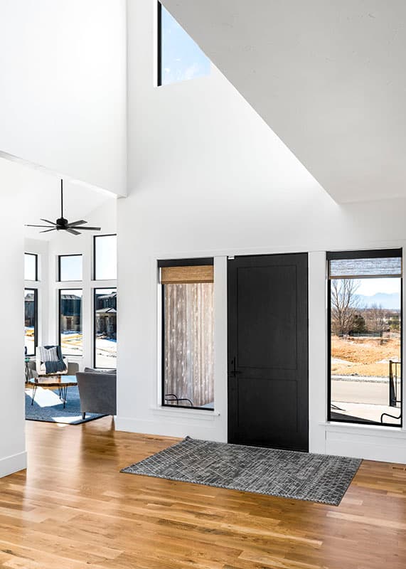 Modern house interior featuring a black paneled door, tall windows, vaulted ceiling with a fan, and an open view to a living area.