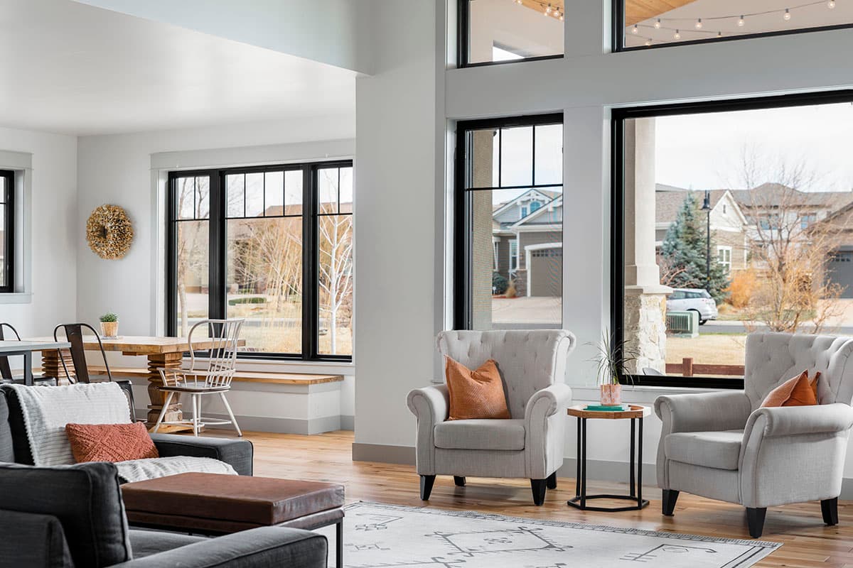 Interior view of a living room with a dining area, featuring large black-framed windows, a wooden table with chairs, and seating.