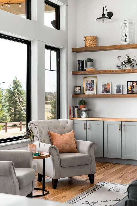 Interior view of a living room with large windows, built-in gray cabinets, wooden floating shelves, and tufted armchairs.