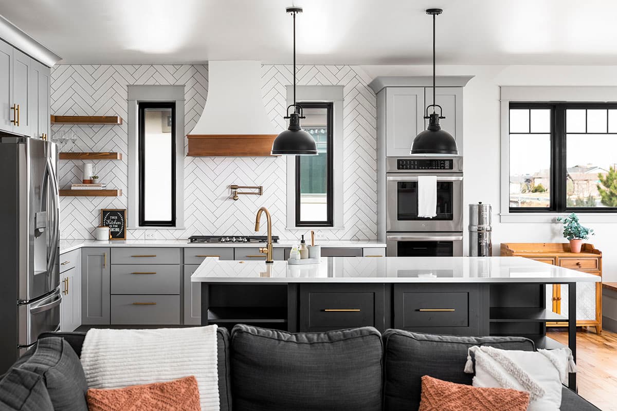 Modern kitchen interior with gray cabinets, white herringbone tile backsplash, a large island with a waterfall countertop, and pendant lighting.