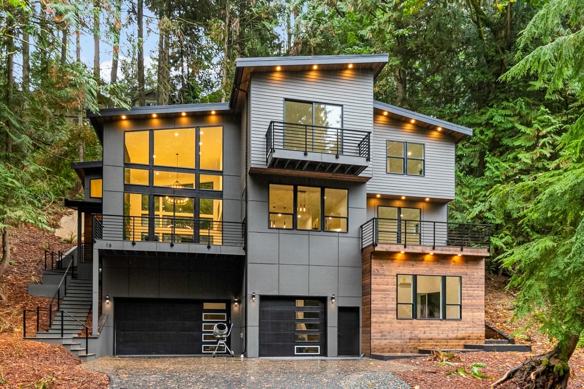 House plan exterior with modern style, two stories, black garage doors, multiple balconies, and large windows. House set amidst trees.