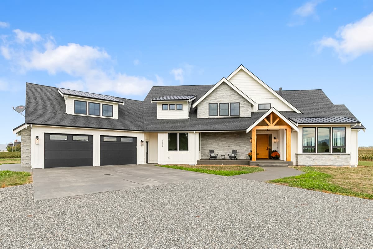 A modern farmhouse with a grey stone facade, black garage doors, and a welcoming front porch. Blue sky and gravel driveway complete the scene.