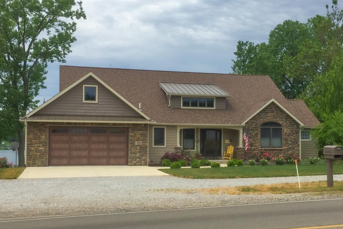 House plan exterior. One-story house with gabled roof, stone facade, and attached garage. Includes a dormer with metal roof.