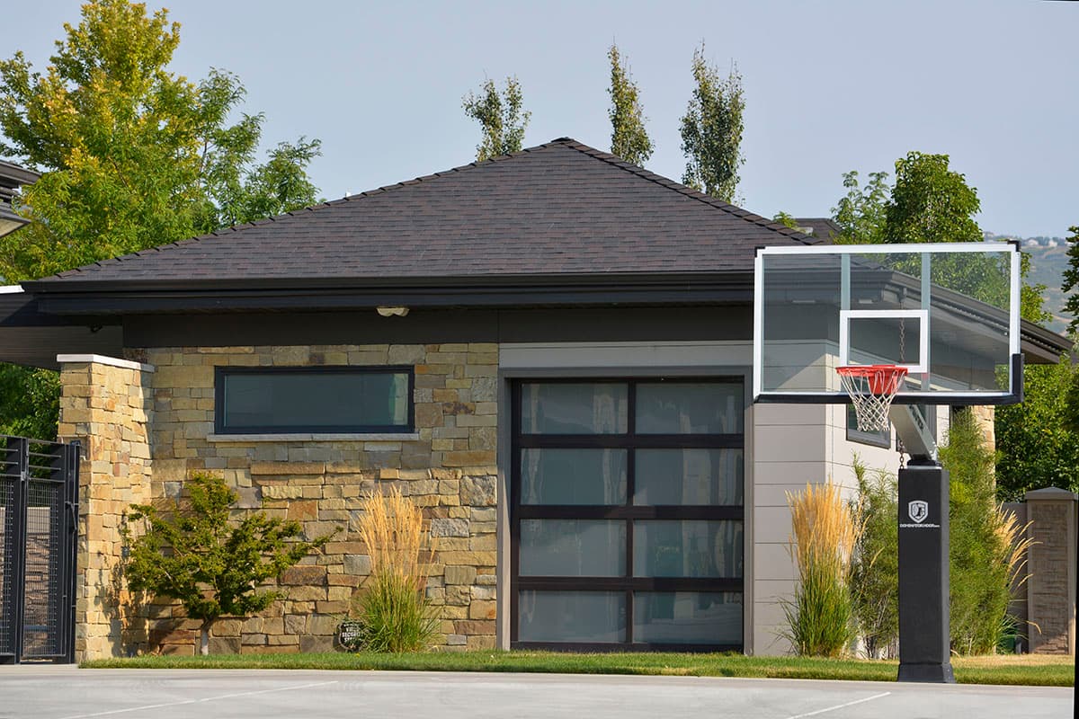House plan exterior. One-story structure with a gable roof. Features include stone veneer, glass garage door, and a basketball hoop.