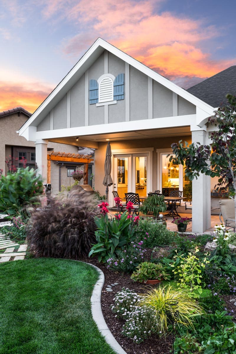 House plan exterior with gabled roof, decorative gable vent, covered patio with outdoor seating, and landscaped yard.