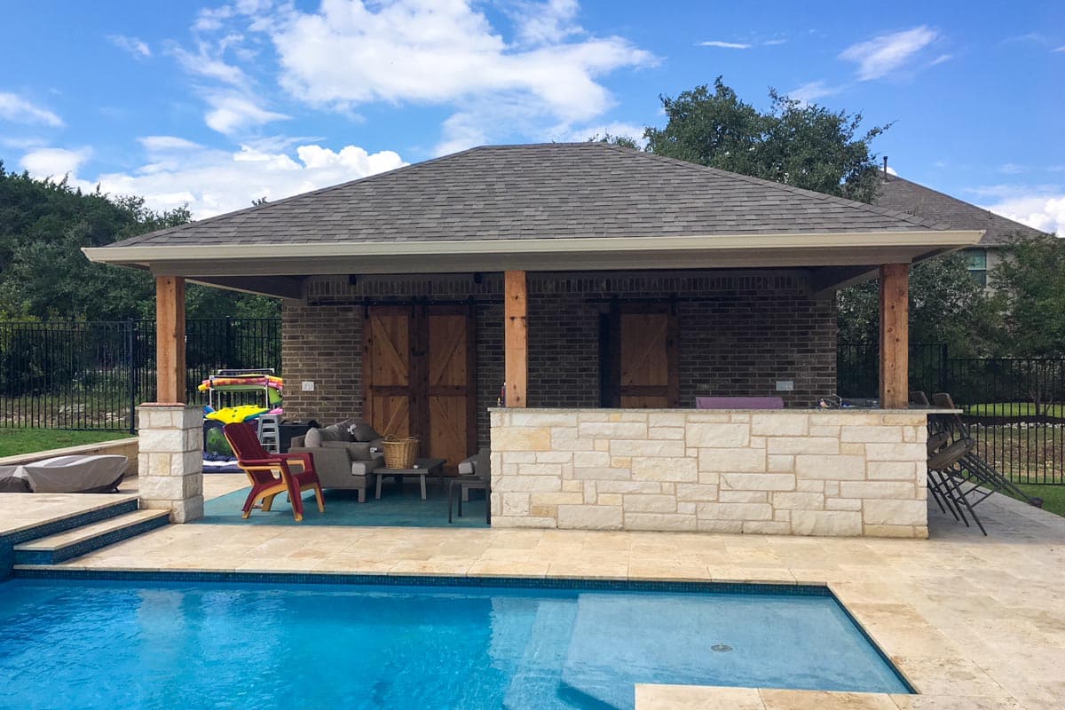 Covered outdoor living space with brick exterior, wood sliding doors, stone bar, and adjacent swimming pool.