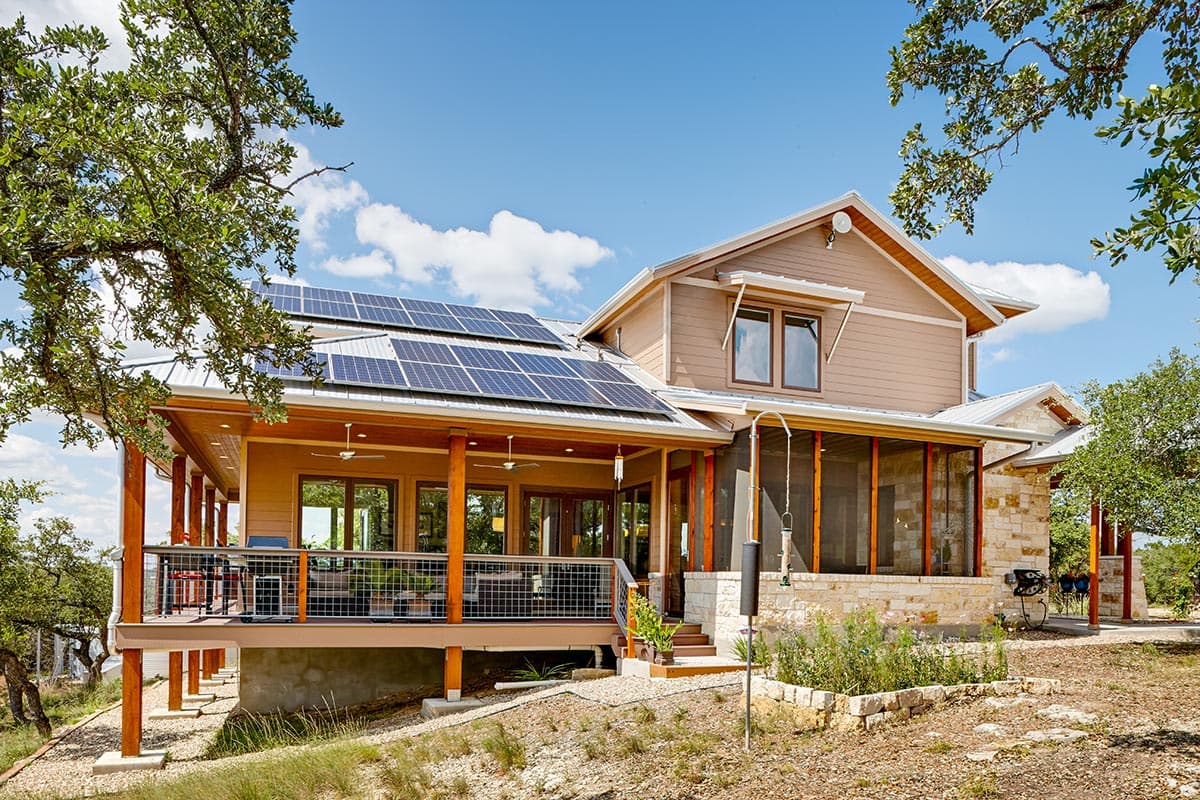 House plan exterior with solar panels, wraparound porch, and stone accents. Two stories with gabled rooflines.