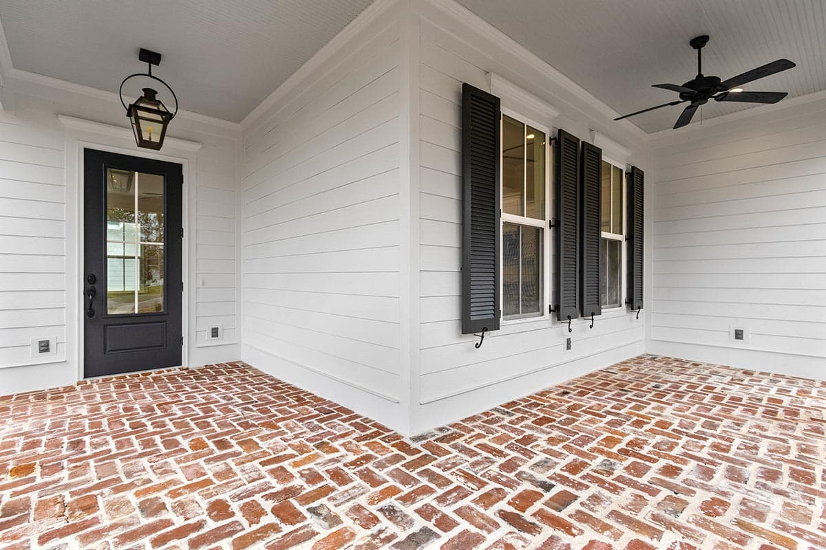 House plan exterior with a covered porch, white lap siding, black shutters, paneled door, and herringbone brick flooring.