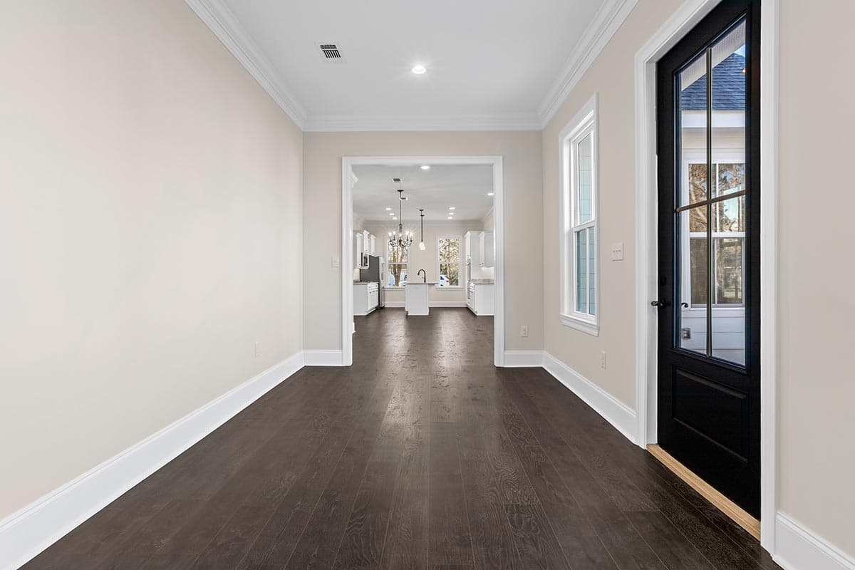 Interior view of a home showing dark wood floors, a black paneled door with glass, and an open view into a kitchen with white cabinets and island.