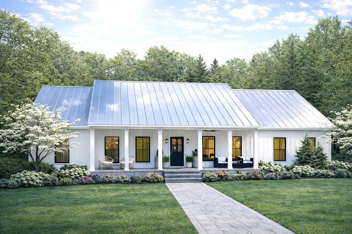 A white farmhouse with a metal roof is framed by lush green trees. A paved walkway leads to the front door, with porch seating visible.