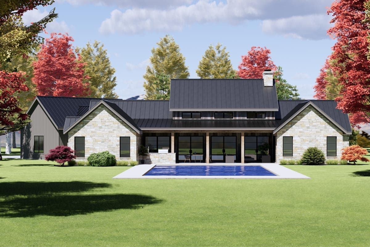 A modern house with a black roof and stone facade. It has a pool in front, set against a backdrop of trees with fall foliage.