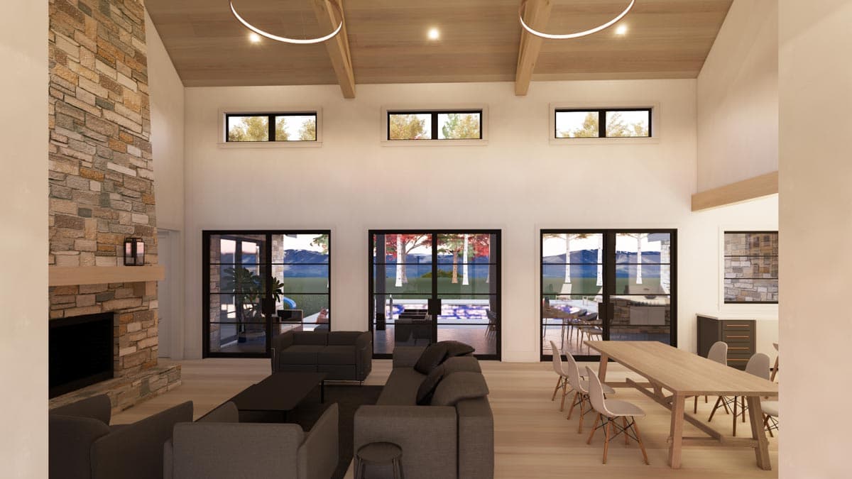 A modern living room with a stone fireplace, large windows overlooking a scenic backyard, and a long dining table.