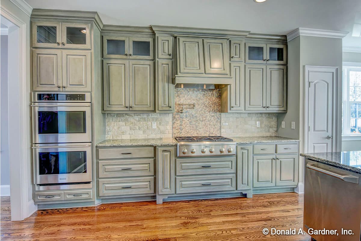 Kitchen with stacked double ovens, gas range, custom gray cabinetry with glass upper doors, and granite countertops.