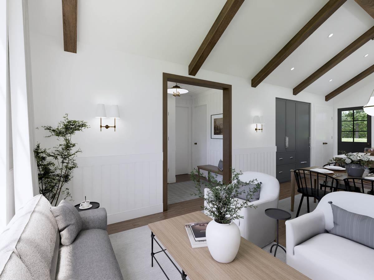 A bright living space featuring exposed wooden beams, white walls, and cozy furniture. A doorway leads to a dining area with a table set for a meal.