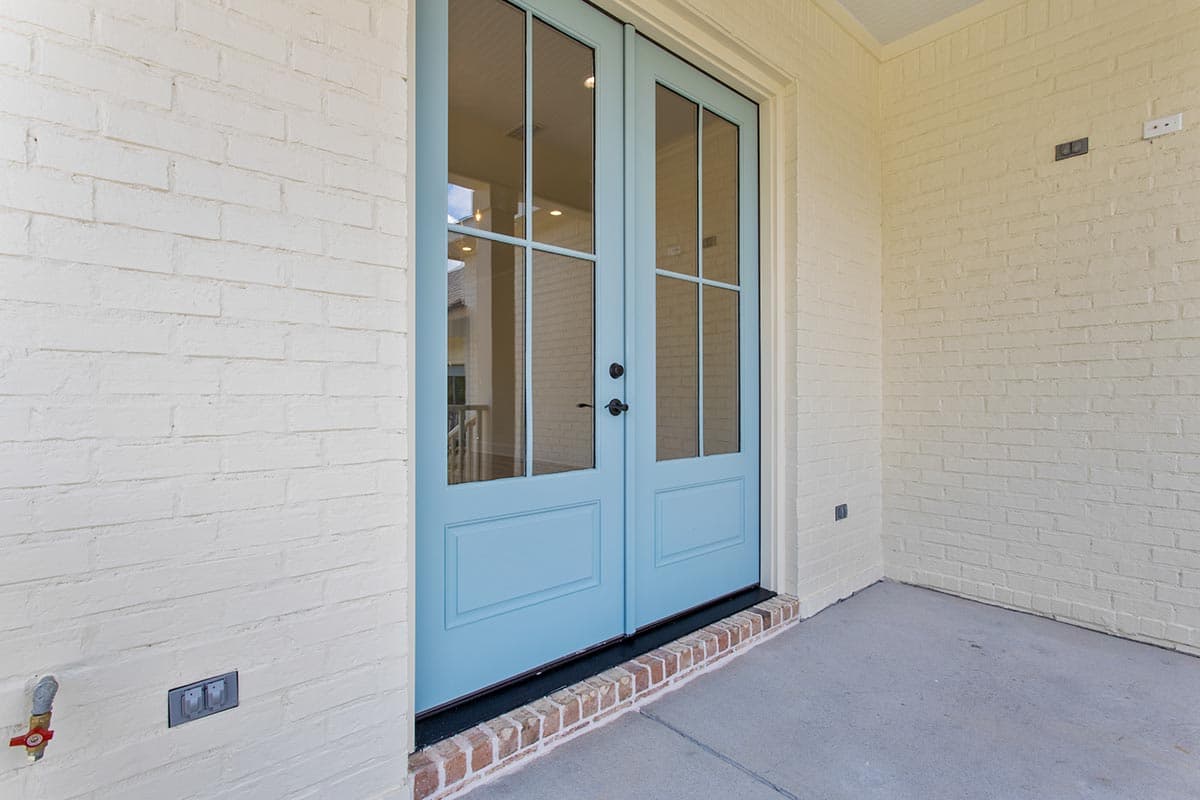 House plan exterior: light brick walls, double glass doors with light blue panes and trim, brick step, concrete porch.
