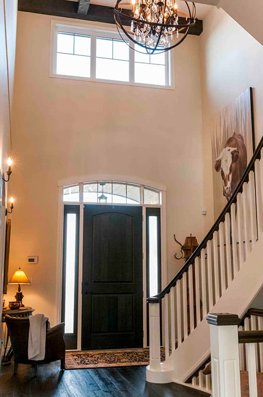 Interior view of a two-story foyer with a dark wood door, transom window, chandelier, and staircase.